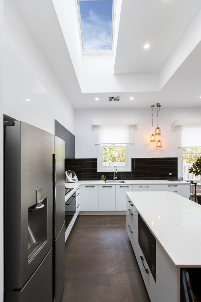 Skylight above a galley-style kitchen with black tile splashback, dark cabinetry, and glass pendant lights. Benefits of daylighting